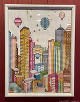 Full view of framed cross stitch art depicting tall colorful town homes and several hot air balloons floating in the sky, with a white frame and glass cover.