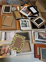 Multiple picture frames stacked on a surface showing a variety of styles including wood, metal, and patterned frames in assorted sizes.