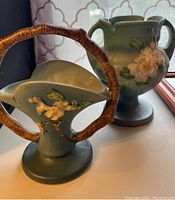 Photo showing Roseville Apple Blossom basket with a round faux wicker handle and matching vase in blue-green glaze with white apple blossom flowers.