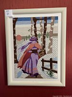 Full view of the framed needlepoint tapestry showing a woman walking in the snow with birch trees and a fence on a red wall.