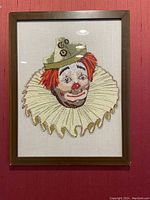 Framed Sad Clown crewel embroidery artwork with clown face wearing a green hat and ruffled collar, hung on red wall.