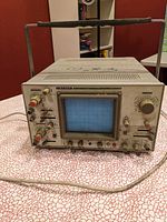 Leader LBO-523 oscilloscope showing CRT screen, control knobs, input connectors, metal casing with handle, placed on patterned surface indoors.