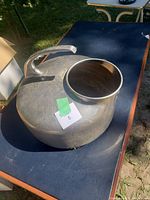 Full view of antique stainless steel milk can with handle and lid, placed on a blue table outdoors.