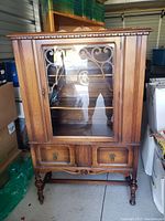 Front view of antique walnut china cabinet showing glass door with decorative wood overlay, drawers below, and turned legs on caster wheels.