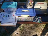 Overall view of table showing multiple plastic containers filled with small bottles of acrylic paint, paint palette with mixed colors, and a metal storage case.