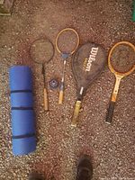 Photo showing four rackets, one in a Wilson cover, and a blue rolled yoga mat on gravel.