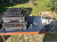 Multiple VHS and DVD players stacked on a table outdoors under sunlight, with various power cords.