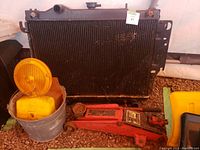 Radiator, red hydraulic car jack, caution light in metal bucket on gravel ground