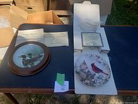 Two collectible plates displayed on a table, one round and framed with a loon image, the other square with cardinals, both with certificates of authentication visible.