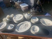 Photo of entire Kutani Japan hand painted porcelain dinnerware set arranged on table showing multiple plates, bowls, serving platter, teapot, sugar bowl, teacups and saucers with traditional Japanese landscape and scenery motifs.