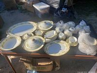 Full china service set laid out showing platters, serving dishes with lids, bowls, cups, and plates wrapped in bubble wrap or arranged on table with clear floral and gold accent pattern.