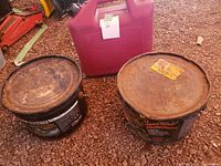 Two buckets of Black Gold roofing tar and a red gas can on dirt ground.