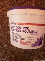 White 2L bucket of CGC Sheetrock Dust Control Drywall Compound with purple and white label on reddish gravel.
