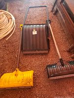 Three shovels: two with wide yellow plastic blades and one black blade shovel with metal frame handle, shown on gravel surface.
