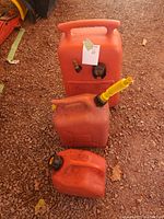 Top view showing all three orange gas cans of varying sizes on dirt ground. Each has unique caps and some have yellow spouts attached. Signs of wear visible.