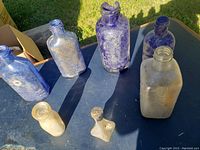 Seven vintage bottles arranged on a table outside, showing dirt and residue, in cobalt blue and clear glass.