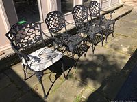 Four cast aluminum chairs on stone patio, leftmost with cushion