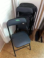 Set of eight black metal folding chairs stacked against a wall with one chair unfolded in front showing seat and back