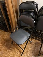 Side angled view showing a folding chair with black upholstered seat and curved backrest, next to a stack of similar chairs.
