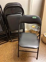 Six black metal folding chairs stacked with one chair unfolded in front showing padded upholstered seat and backrest.