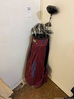 Full view of maroon Wilson golf bag containing golf clubs standing against wall on floor