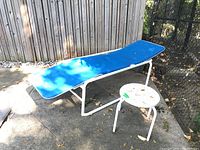 Photo of one outdoor lounge chair with a blue cushion and a round white side table on a concrete patio next to a wooden fence.