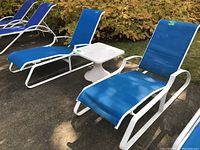 Two blue mesh outdoor lounge chairs with white aluminum frames and one white square fiberglass pedestal table outside on a patio with foliage in background.