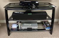 Front view of media stand holding electronic devices (not included) showing three glass shelves and black metal frame.