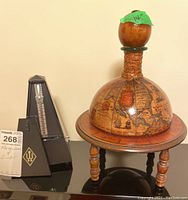Photo showing Wittner metronome with black wood grain plastic housing next to the antiqued globe shaped decanter on its wooden stand, labeled with measurements.