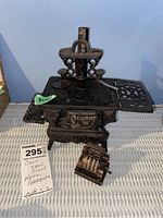Miniature black cast iron stove with 'Crescent' on front and dark brown novelty pencil sharpener shaped like cash register on gray wicker surface against light blue wall.