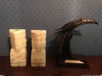 Front and side view of two onyx stone bookends carved as stylized human faces, and a painted wood bird figurine on a wooden base.