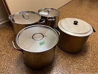 Four stock pots displayed together on floor, three stainless steel with matching stainless lids and one aluminum with a different lid.