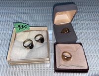 All four rings displayed in individual boxes: two silver-tone with black stones on white cushion, two gold-tone with monogram on grey and pink cushions.