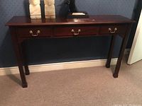 Front view of dark stained wood sofa table showing three drawers and brass-tone bail pulls against a blue wall.