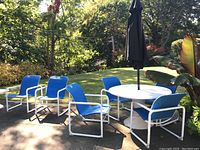 Outdoor setting showing the full set including round white fiberglass table, six blue sling chairs with white aluminum frames, and black umbrella closed in the center of the table.