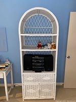 Front view of tall white wicker cabinet with three shelves, one drawer, and cupboard section. Contains some items on shelves not included in sale.