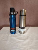 Front view of two Thermos brand thermoses, one blue and one stainless steel, both standing upright on white surface.