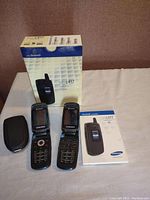 Two Samsung SGH-C417 flip phones, box, manual, and protective case arranged on a beige surface with a brown background.