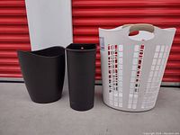Photo showing two black garbage bins and one white plastic laundry hamper in front of a red storage door.
