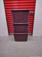 Three stacked red Sterilite tote bins with lids in front of a red storage unit door. Bins appear in good condition with no visible damage.