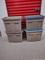 Four Rubbermaid Roughneck tote bins stacked in front of a red storage locker door. Bins are tan colored and lids are two mismatched colors (blue and gray) and two tan. Some writing visible on bins and lids.