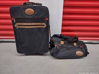 Two-piece Buxton luggage set with black fabric and brown accents, including suitcase and duffle bag on a concrete floor in front of a red wall.