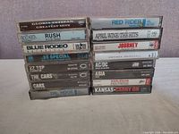 Photo showing front and spine of 16 cassette tapes in clear plastic cases stacked in two piles. Visible titles include Gloria Estefan Greatest Hits, Rush Fly By Night, Anthem, Blue Rodeo Just Like You, .38 Special Wild-Eyed Southern Boys, ZZ Top Eliminator, The Cars, Dr. Hook, Red Rider Neruda, April Wine The Hits, Journey, Amanda Marshall, AC/DC Back In Black, Asia, Harlequin, Kansas Carry On.