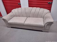 Front angled view of beige sofa showing scalloped, fan-patterned backrest, two seat cushions, and rolled arms.
