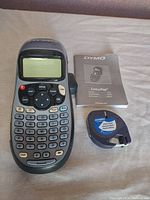 Dymo LetraTag label maker alongside user manual and one printer roll on light fabric surface.
