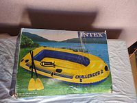 Photo of the Intex Challenger 2 inflatable kayak box showing an image of the yellow and blue kayak with two yellow paddles and a hand pump on a grassy outdoor background near water.