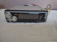 Front view of the JVC car stereo showing control knobs, buttons, digital display, and logo.