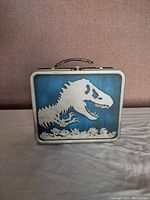 Front side of the metal lunch box showing white T-Rex skeleton silhouette on blue background with black handle on top.
