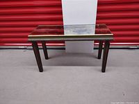 Front view of mid century modern marbled glass topped rectangular stool or small console table with metal trim and wooden legs on concrete floor with red door background.