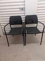 Two black outdoor chairs with woven seats and backrests, metal tubular frames, arms show wear, placed side by side on concrete floor.
